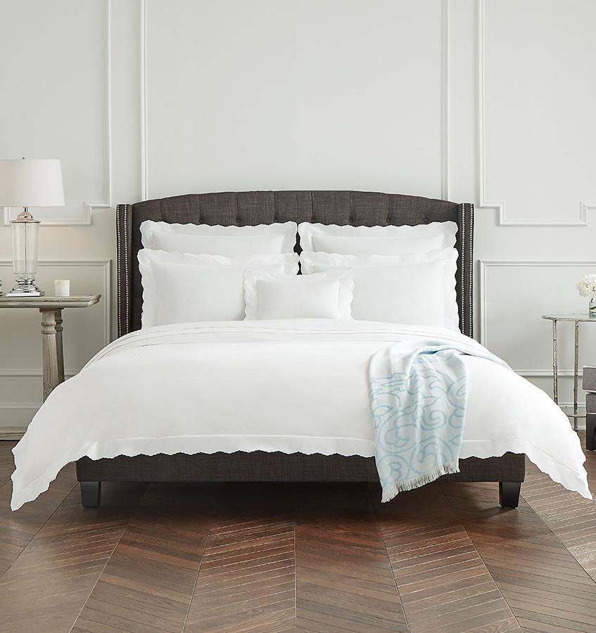 A brown bed in a white bedroom with white SFERRA Pettine bedding with scalloped edges.