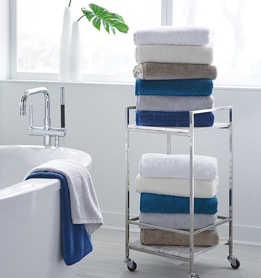 A bathroom with stacks of SFERRA Sarma bath towels in white, ivory and shades of blue.