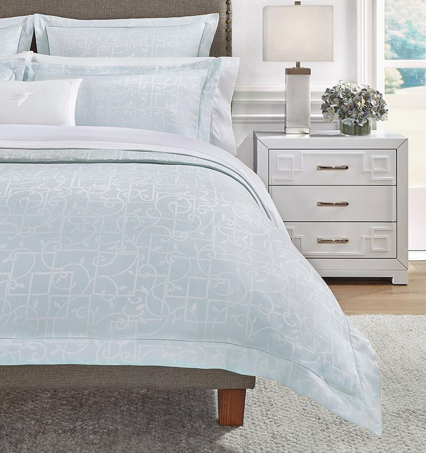 The left side of a bed with a light blue SFERRA Graticcio Duvet Cover woven with a white trellis pattern with matching shams.