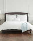 A brown bed in a white bedroom with white SFERRA Pettine bedding with scalloped edges.