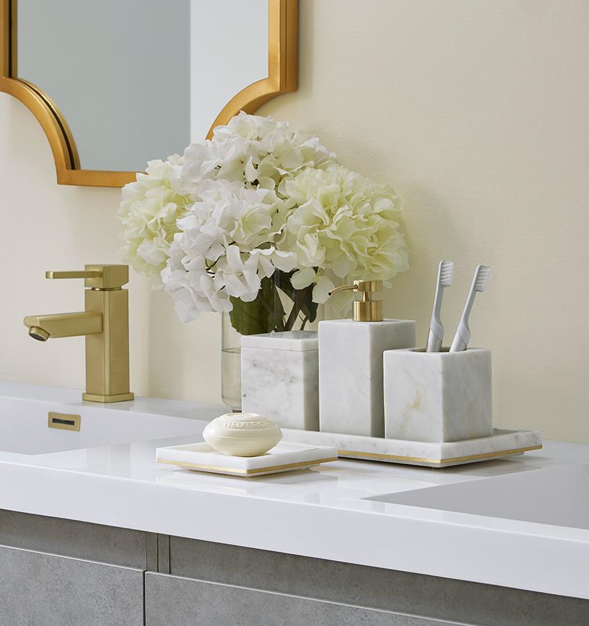 A bathroom counter with gold-trimmed marble SFERRA Pietra bathroom accessories and white hydrangeas.