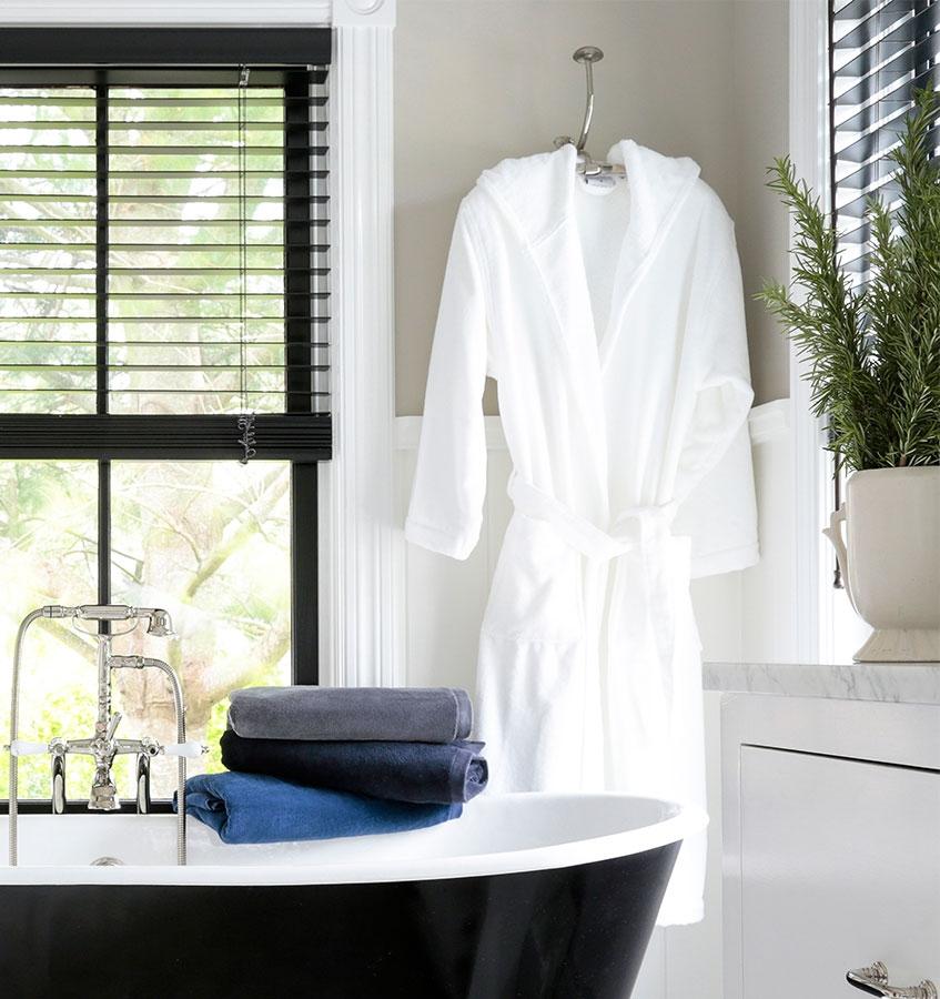 A bathroom with SFERRA Canedo towels stacked on the side of the bath tub and a robe hanging behind it.