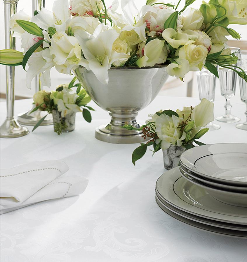 Dishes stacked on a table with a SFERRA Acanthus Tablecloth under them. A vase of white flowers sits behind it.
