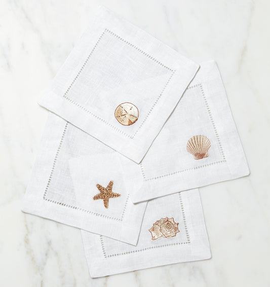 SFERRA Beachcomber cocktail napkins feature four delightful seashell embroideries in golden hues on hemstitched linen napkins.