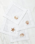 SFERRA Beachcomber cocktail napkins feature four delightful seashell embroideries in golden hues on hemstitched linen napkins.