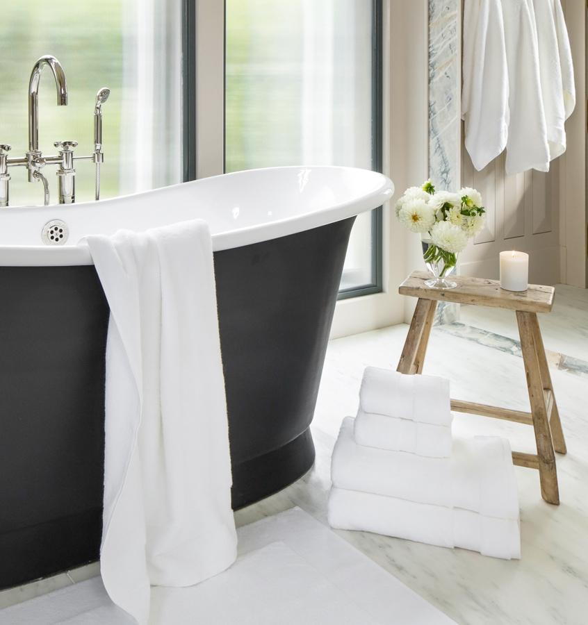 A bathroom with white SFERRA luxury bath towels draped across the bath rub and stacked on the floor.